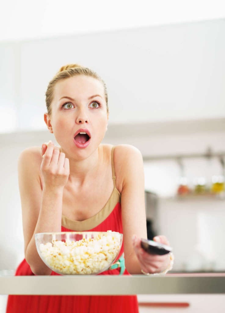 Surprised Young Woman Eating Popcorn And Watching Tv In Kitchen