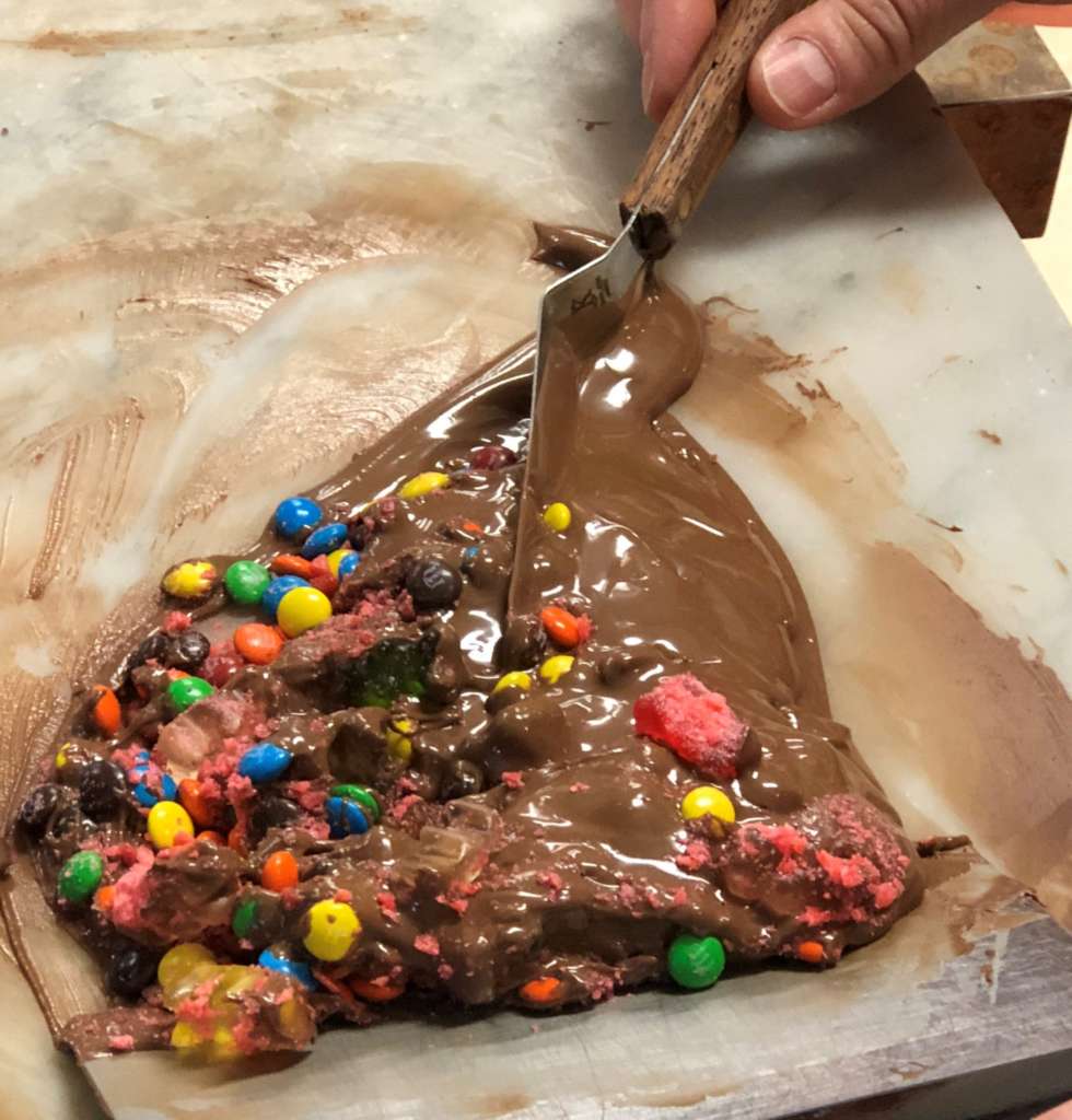A mound of melted chocolate on a marble slab with blue, pink, yellow and green candies being mixed in.