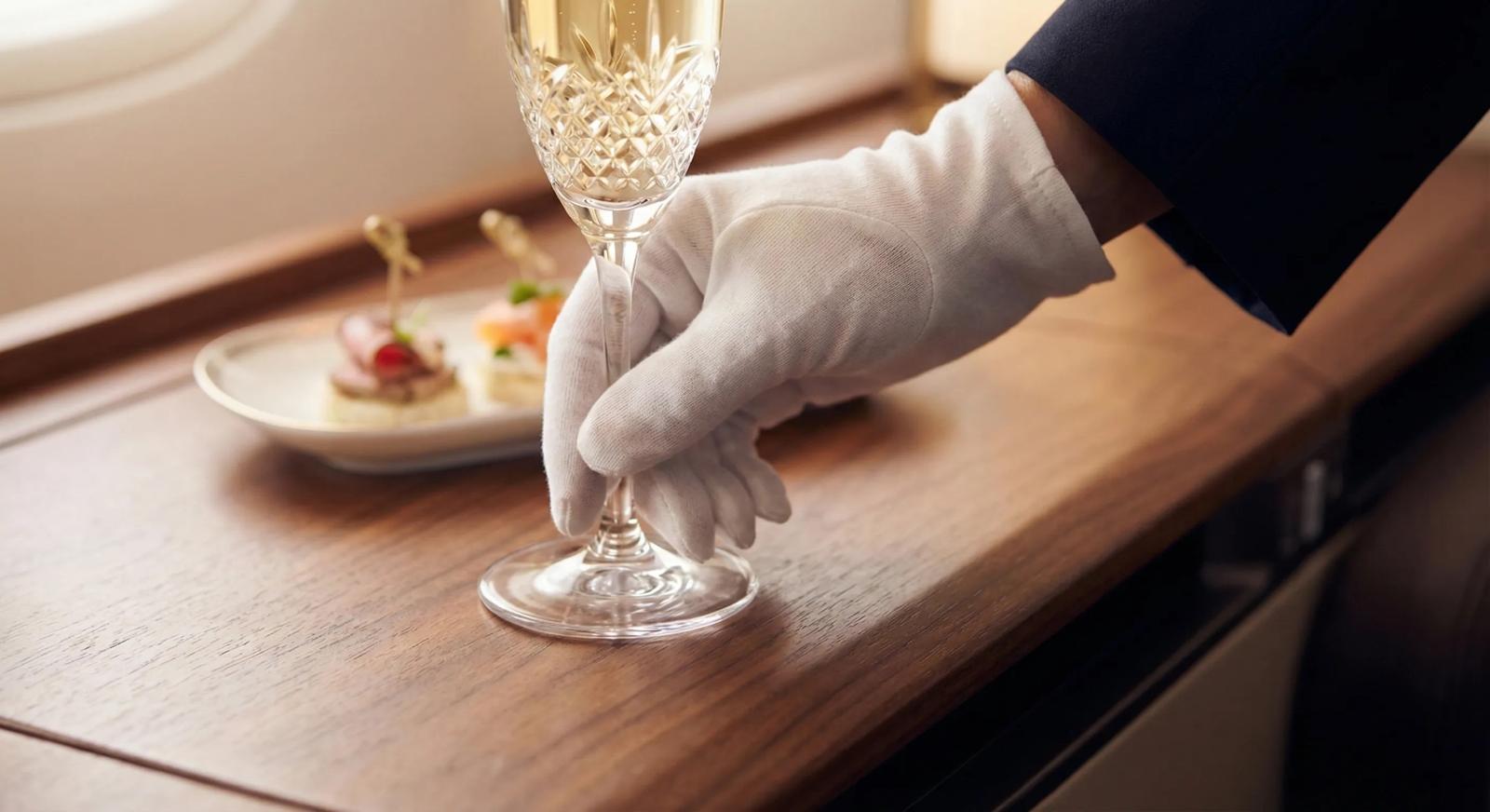 Close-up detail of a flight attendant in a white glove placing a glass of champagne on a wooden table, emphasizing premium service